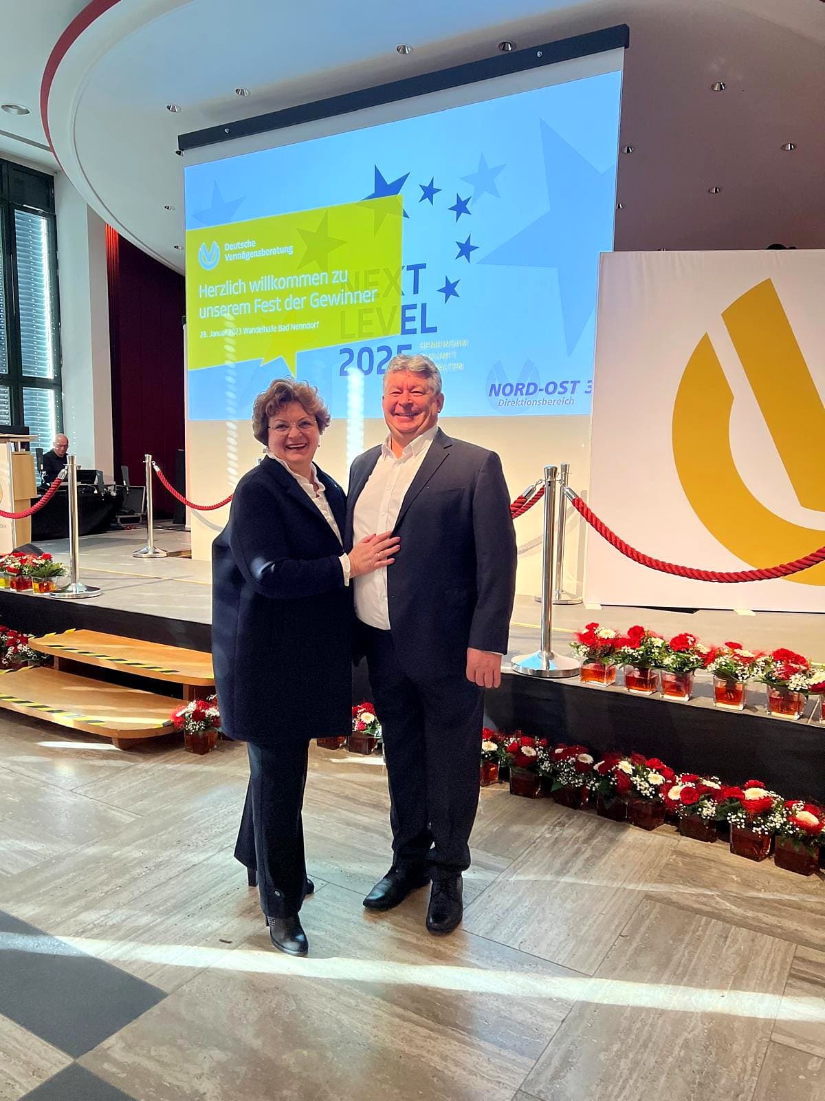 Larissa Etzel und ein Begleiter im Festsaal vor einer Leinwand der Deutschen Vermögensberatung.